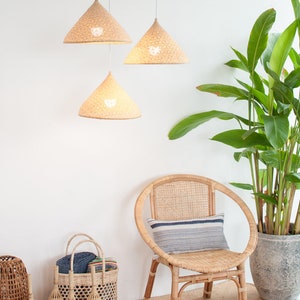 May include: Three woven conical pendant lights hang from the ceiling in a white room. A wicker chair with a striped cushion sits in the corner, next to a potted plant. Several woven baskets are on the floor.