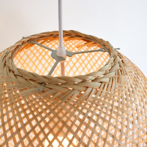 Op de afbeelding: Een geweven bamboe hanglamp met een wit snoer. De lamp is rond en heeft een geweven patroon.