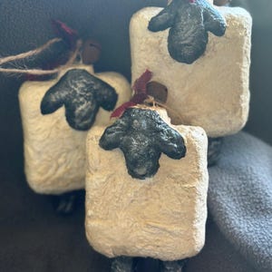May include: Three handmade sheep ornaments. Each has a rectangular body with a textured, off-white surface. The sheep have black faces and legs, with a small red bow tied around their necks. They are strung with twine for hanging.