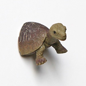 Könnte beinhalten: Kleine Schildkrötenfigur aus Kunststoff. Die Schildkröte hat einen braunen und beigefarbenen Panzer mit einem strukturierten Muster. Kopf und Beine sind hellbeige. Die Figur steht vor einem schlichten weißen Hintergrund.