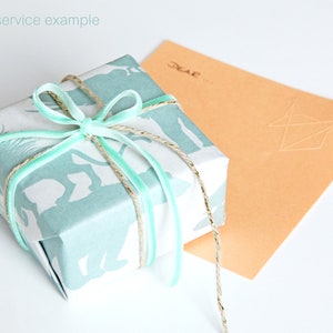 Puede incluir: Una caja de regalo azul claro con un dise&ntilde;o de estampado de animales blancos est&aacute; envuelta con una cinta azul claro y un cordel. La caja est&aacute; sobre una superficie blanca junto a un sobre de color melocot&oacute;n con la palabra "Querido..." escrita en &eacute;l.