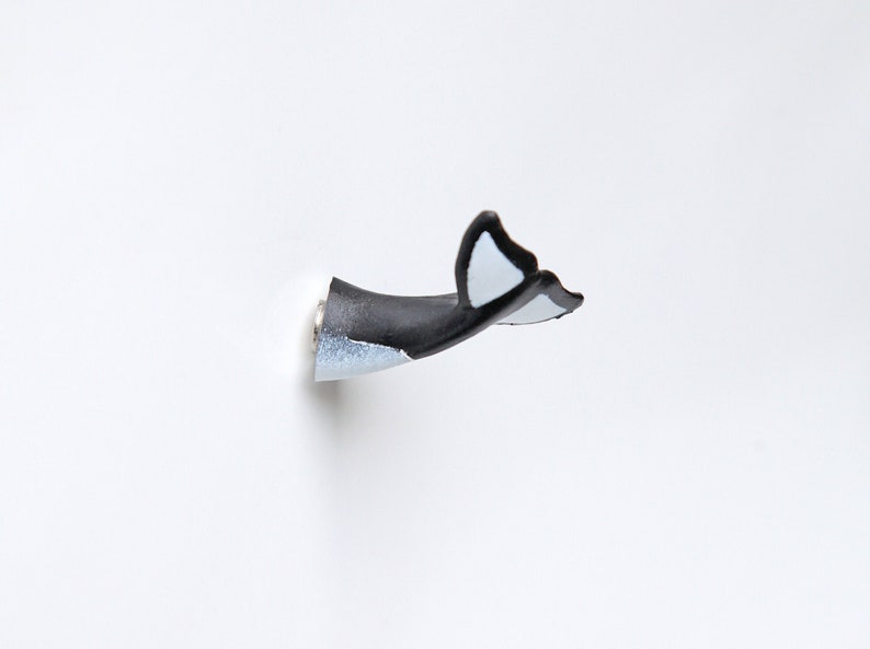 Puede incluir: Un gancho de pared de cola de ballena negro y blanco. El gancho est&aacute; hecho de pl&aacute;stico y tiene un tornillo plateado.