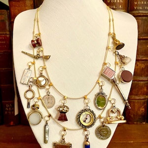 May include: A gold-toned necklace with multiple charms of various shapes and sizes, including keys, lockets, and compasses. The charms are suspended from delicate chains. The necklace is displayed on a white mannequin bust against a backdrop of antique books.
