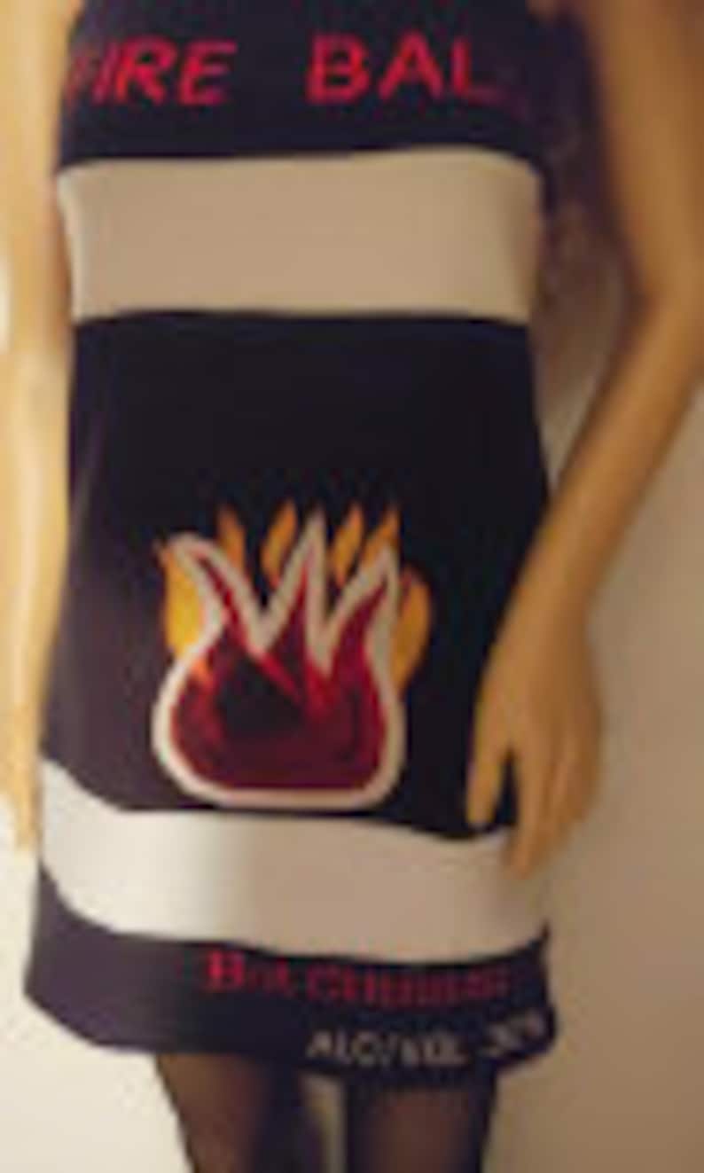May include: A black and white striped dress with a pink "FIRE BALL" logo and a red and orange flame graphic. The dress has a white stripe at the bottom with the text "THE ORIGINAL" and "ALCOHOL 12%".