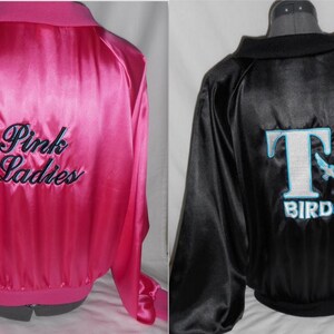 Puede incluir: Dos chaquetas de sat&eacute;n, una rosa con "Pink Ladies" bordado en la espalda, y una negra con un logotipo "T Birds" blanco con un p&aacute;jaro azul.