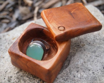 May include: A wooden ring box with a hinged lid, open to reveal a ring with a light blue gemstone. The box is made of a light brown wood with a smooth finish.