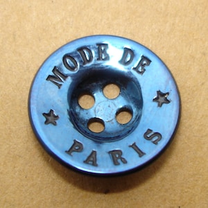 May include: A blue metal button with the words "MODE DE PARIS" engraved around the edge. The button has four holes for sewing.