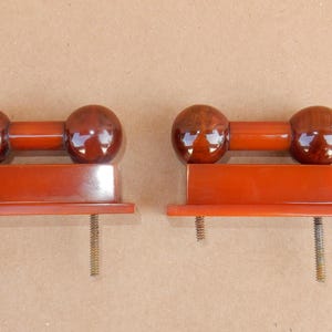 May include: Two vintage, reddish-brown cabinet knobs. Each knob features a pair of glossy, spherical ends connected by a cylindrical bar, mounted on a rectangular base with a threaded screw. The knobs are likely made of Bakelite or a similar material.