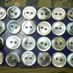 May include: A collection of 24 iridescent mother-of-pearl buttons in a blue box. The buttons are round and have two holes.