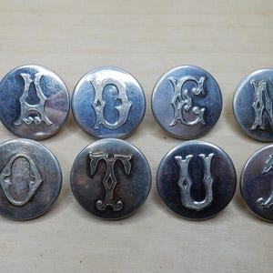 May include: Eight silver-toned metal buttons with different letters of the alphabet, each letter is in a decorative font.