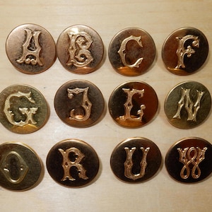 Könnte beinhalten: Zwölf goldfarbene Metallknöpfe, jeder mit einem anderen Buchstaben des Alphabets geprägt.