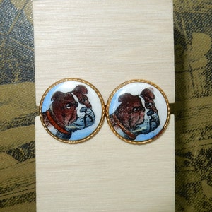 May include: A pair of cufflinks featuring a white and brown bulldog head on a blue and white background. The cufflinks are gold-toned and have a round shape.