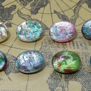 Può includere: Otto cabochon in vetro dicroico in vari colori, tra cui verde, rosa e blu, con effetti iridescenti. I cabochon sono rotondi e poggiano su uno sfondo a forma di mappa.