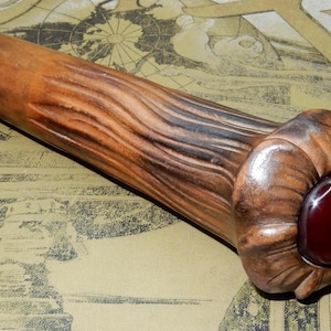 Könnte beinhalten: Ein handgefertigter Flaschenverschluss aus Holz mit einem geschnitzten, strukturierten Design. Der Verschluss hat einen dunkelroten, abgerundeten Edelstein am Ende. Das Holz hat eine warme, braune Farbe mit dunkleren Streifen.