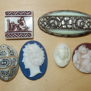 Könnte beinhalten: Sortiment von Vintage-Cameo-Broschen mit verschiedenen Designs, darunter eine Szene mit Figuren, ein Profilporträt und ein florales Motiv. Die Broschen sind aus weißer und blauer Porzellan mit goldenen Akzenten.
