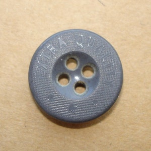 Op de afbeelding: Een grijze plastic knoop met vier gaten en de tekst "EXTRA QUALITY" gegraveerd rond de rand.