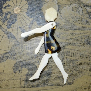 Puede incluir: Una muñeca de madera vintage con extremidades articuladas. La muñeca tiene cabeza y extremidades de color claro, con un torso oscuro y estampado. La muñeca está posada en posición de caminar sobre un fondo de mapa vintage.