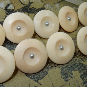 Puede incluir: Diez botones redondos de color crema con un centro plateado. Los botones tienen un diseño circular en relieve.