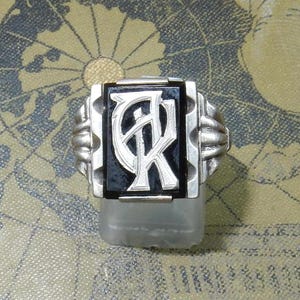 Könnte beinhalten: Ein silberner Ring mit einem schwarzen Onyxstein, der in einem rechteckigen Rahmen gefasst ist. Der Stein trägt ein silbernes Monogramm der Buchstaben "AR".