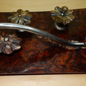 May include: A decorative tray with a silver metal design in the shape of a branch with three carved wooden flower accents. The tray is made of dark wood with a marbled pattern.