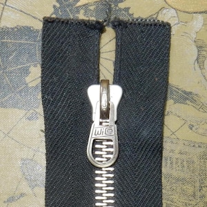 May include: Close-up of a black zip fastener with a silver pull tab. The zip is set against a patterned fabric with a map design in shades of tan and blue. The pull tab has the letters "WIC" on it.