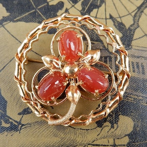 May include: A gold-toned brooch featuring a three-dimensional flower design with three red gemstone cabochons. The flower is set within a circular frame of twisted wire.