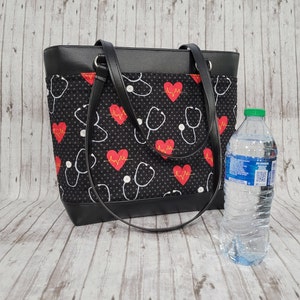 May include: Black tote bag with a black and white pattern of hearts and stethoscopes. The bag has a black leather trim and two black handles. A clear plastic water bottle is next to the bag.