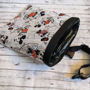 May include: A white fabric bag with a black strap and a Mickey Mouse and Minnie Mouse print. The bag has a black zipper and a clear plastic lining.