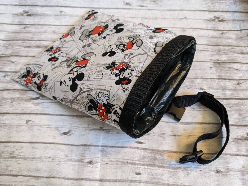 May include: A white fabric bag with a black strap and a Mickey Mouse and Minnie Mouse print. The bag has a black zipper and a clear plastic lining.