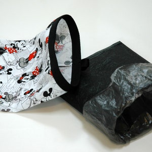 May include: A white fabric bag with a black trim and a Mickey Mouse print. The bag is folded in half and is lying on a white surface. Next to the bag is a black mesh bag with a black plastic bag inside.