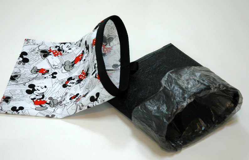 May include: A white fabric bag with a black trim and a Mickey Mouse print. The bag is folded in half and is lying on a white surface. Next to the bag is a black mesh bag with a black plastic bag inside.