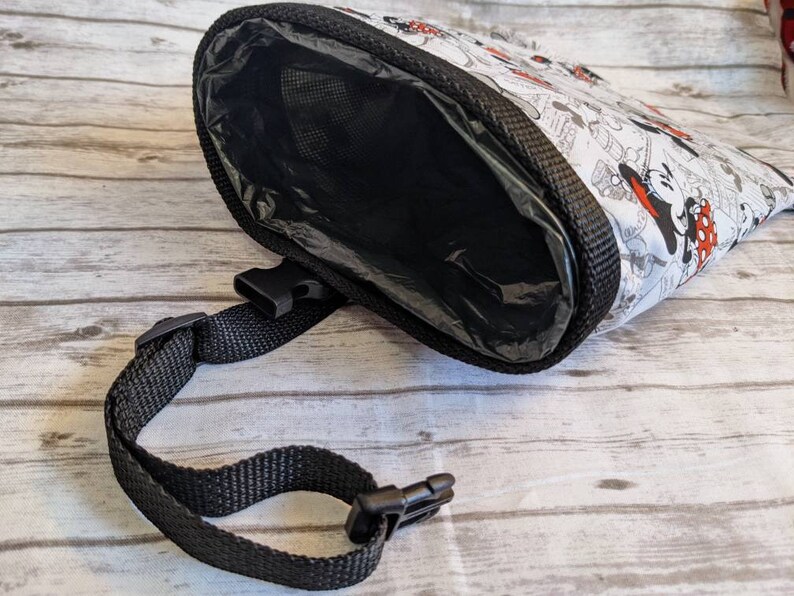 May include: A black and white dog poop bag dispenser with a Mickey Mouse and Minnie Mouse print. The dispenser has a black strap with a buckle.