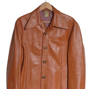 May include: A vintage brown leather jacket with a button-up front and a wide collar. The jacket is hanging on a wooden hanger. The leather has a slightly worn appearance, suggesting it is a pre-owned item.