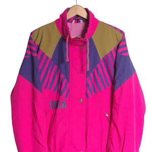 May include: A vintage magenta jacket with a high collar and a drawstring hood. The jacket features colour-blocked panels in brown and purple with diagonal stripes. The word "SILA" is printed on the front. The jacket is hanging on a wooden hanger.