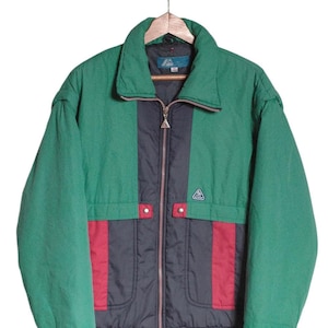 May include: A green, black, and red vintage ski jacket with a zipper closure and a logo on the chest. The jacket has a stand-up collar and a ribbed waistband.