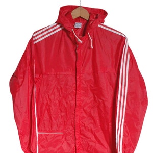 May include: A red hooded jacket with white stripes down the sleeves and sides. The jacket has a zipper closure and a small pocket on the left side. The Adidas logo is visible on the bottom right side of the jacket.