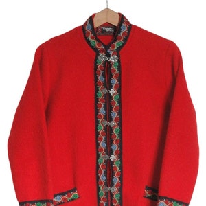 Puede incluir: Una chaqueta de lana roja con cuello alto y ribetes decorativos. Los ribetes presentan un fondo negro con bordados verdes, azules y rojos. La chaqueta tiene botones plateados y la etiqueta "Lapp-Style" es visible en el cuello.