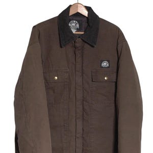 May include: Brown work jacket with a dark gray collar and two chest pockets. The jacket has a button-down front and a small logo patch on the chest. The jacket is hanging on a wooden hanger.