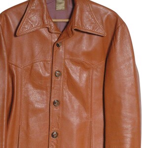 May include: A brown leather jacket with a wide collar and button-up front. The jacket is hanging on a wooden hanger. The leather has a slightly distressed appearance, with visible stitching details and two front pockets.