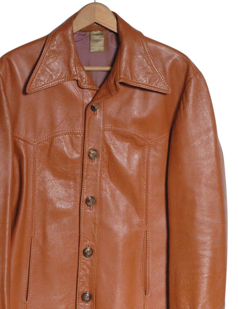 May include: A brown leather jacket with a wide collar and button-up front. The jacket is hanging on a wooden hanger. The leather has a slightly distressed appearance, with visible stitching details and two front pockets.