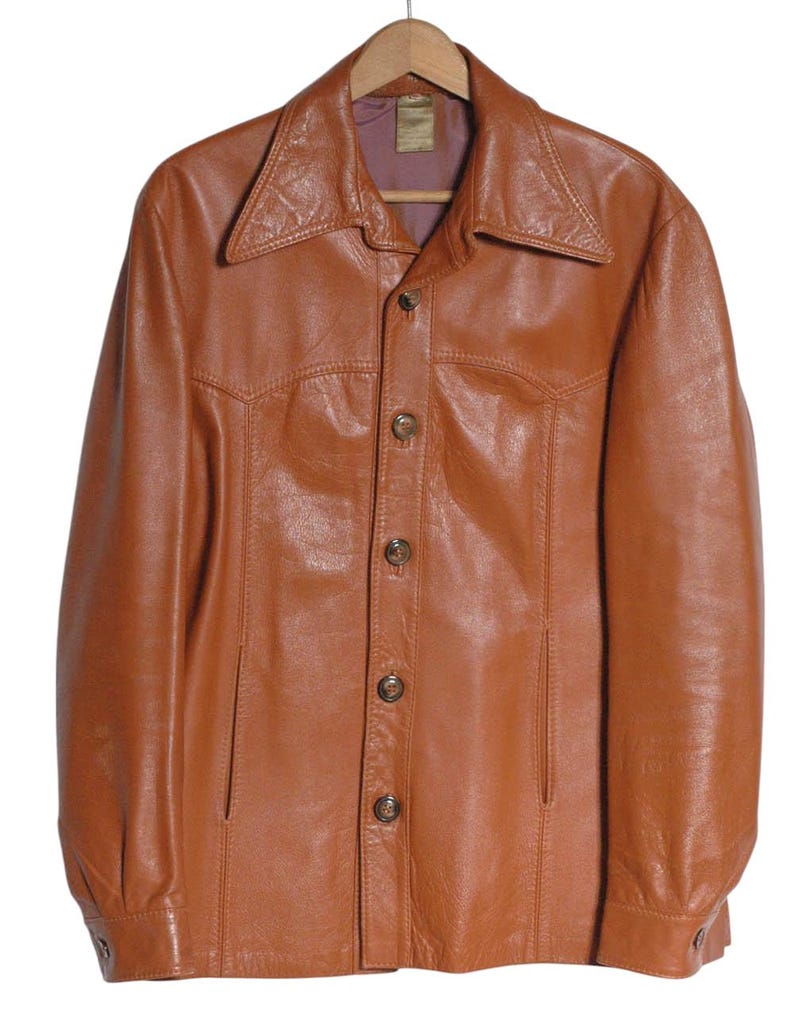 May include: A vintage brown leather jacket with a button-up front and a wide collar. The jacket is hanging on a wooden hanger. The leather has a slightly worn appearance, suggesting it is a pre-owned item.