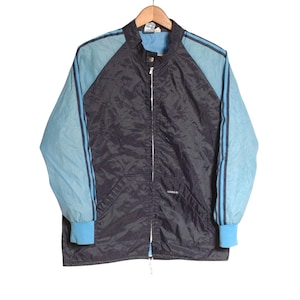 Könnte beinhalten: Vintage Adidas Trainingsjacke in Marineblau und Hellblau. Die Jacke hat hellblaue Ärmel mit zwei marineblauen Streifen, einen marineblauen Körper und einen durchgehenden Reißverschluss. Sie hängt an einem Holzbügel.