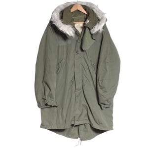 Puede incluir: Parka verde oliva con capucha ribeteada con piel sintética. La chaqueta tiene cintura ajustable con cordón, cierre de cremallera y solapa abotonada. La parka está colgada de una percha de madera.