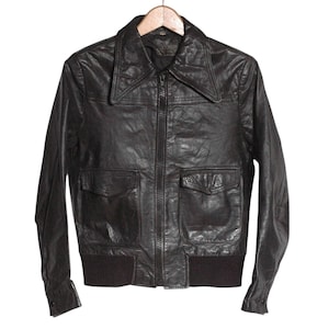 May include: A vintage black leather jacket with a collared neckline, a zipper closure, and two front pockets. The jacket has a ribbed knit hem and cuffs. It is hanging on a wooden hanger against a white background.