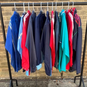 May include: A black metal clothing rack holding a row of various athletic jackets in shades of blue, red, navy, and teal. The jackets feature long sleeves, some with white stripes, and are hung on individual plastic hangers against a brick wall.