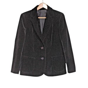 Op de afbeelding: Een zwarte blazer met twee knopen en twee voorzakken, hangend aan een houten hanger. De blazer heeft een klassiek ontwerp en is gemaakt van een zacht, fluwelig materiaal. De jas is effen van kleur.