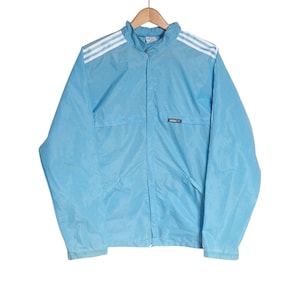 May include: Light blue Adidas track jacket with white stripes on the shoulders. The jacket has a full zipper, two front pockets, and the Adidas logo on the chest. The jacket is hanging on a wooden hanger.