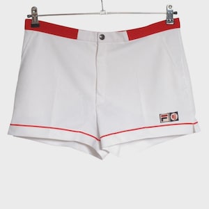 May include: White athletic shorts with red trim and a red waistband. The shorts feature a small logo on the lower right leg. The shorts are hanging on a metal hanger.