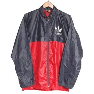 Peut inclure: Un coupe-vent à manches longues présentant un design bicolore avec une partie supérieure bleu marine foncé et une partie inférieure rouge vif. La veste possède une fermeture éclair intégrale, un col montant et le logo trèfle Adidas blanc.