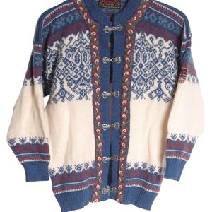 May include: A blue and off-white knit cardigan featuring a complex geometric pattern, long sleeves, and a front closure with four silver-toned metal clasps. The garment includes a decorative trim along the opening and a small patch on the left sleeve.